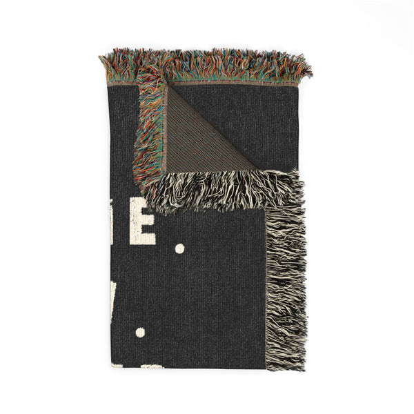 A folded dark charcoal woven throw blanket with multicolor fringe, featuring a large cream letter 'E' and small cream dots on a white background.