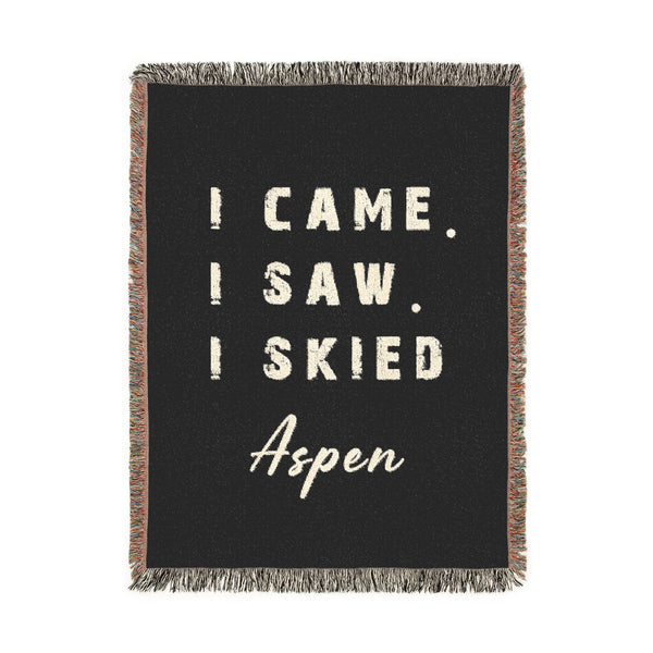 A rectangular black woven throw blanket with fringe edges, featuring the phrase 'I CAME. I SAW. I SKIED ASPEN