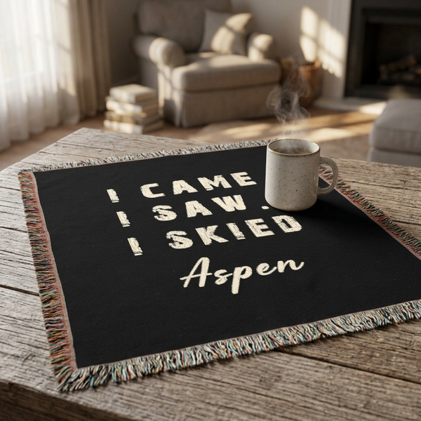 A black woven throw blanket with multicolor fringe and cream text reading 'I CAME I SAW I SKIED Aspen' is draped over a rustic wooden table in a cozy living room setting, with a white speckled mug resting on it.