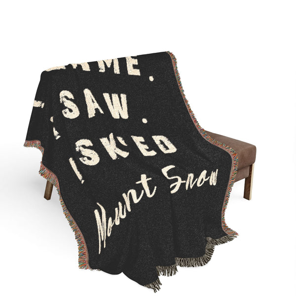 A black woven throw blanket with cream-colored text reading 'I CAME. I SAW I SKIED Mount Snow' draped over a brown armchair against a white background.
