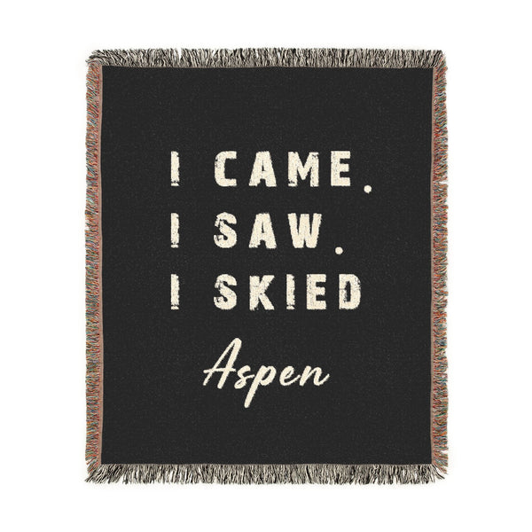 A square black woven throw blanket with fringe edges, featuring the phrase 'I CAME. I SAW. I SKIED ASPEN