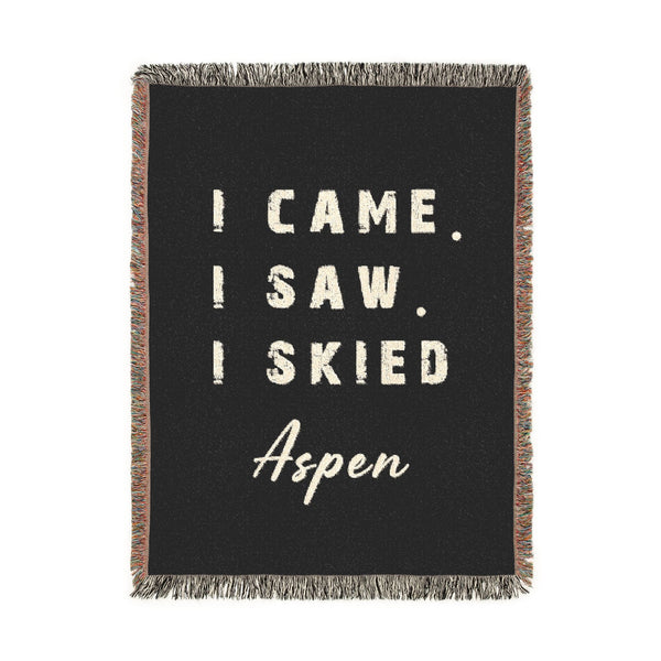 A rectangular black woven throw blanket with fringe on all sides, featuring the phrase 'I CAME. I SAW. I SKIED ASPEN