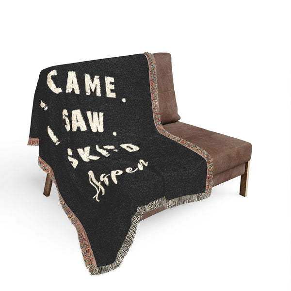 A black woven throw blanket with cream text reading 'I CAME. I SAW. I SKIED ASPEN