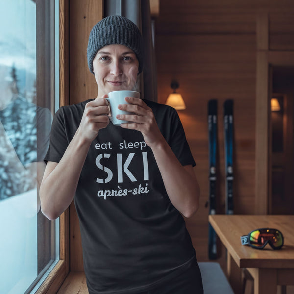 A person wearing a black t-shirt with the text "eat sleep ski après-ski" is standing near a window, holding a cup and smiling.